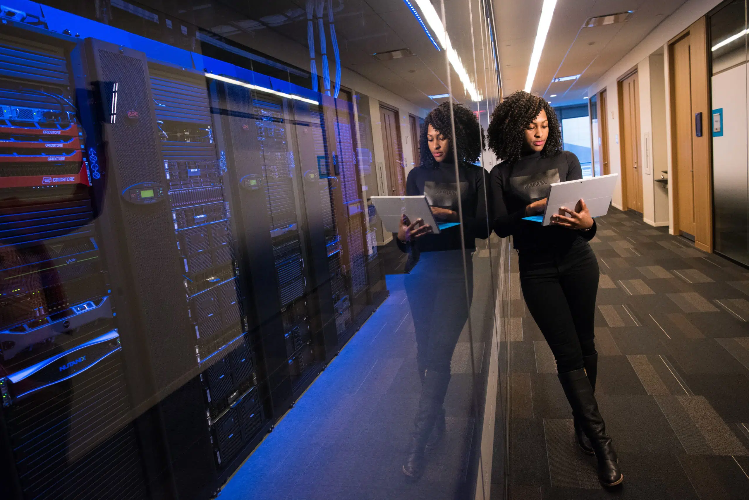Black woman holding a laptop leans into a glass wall where we can see large computer services.