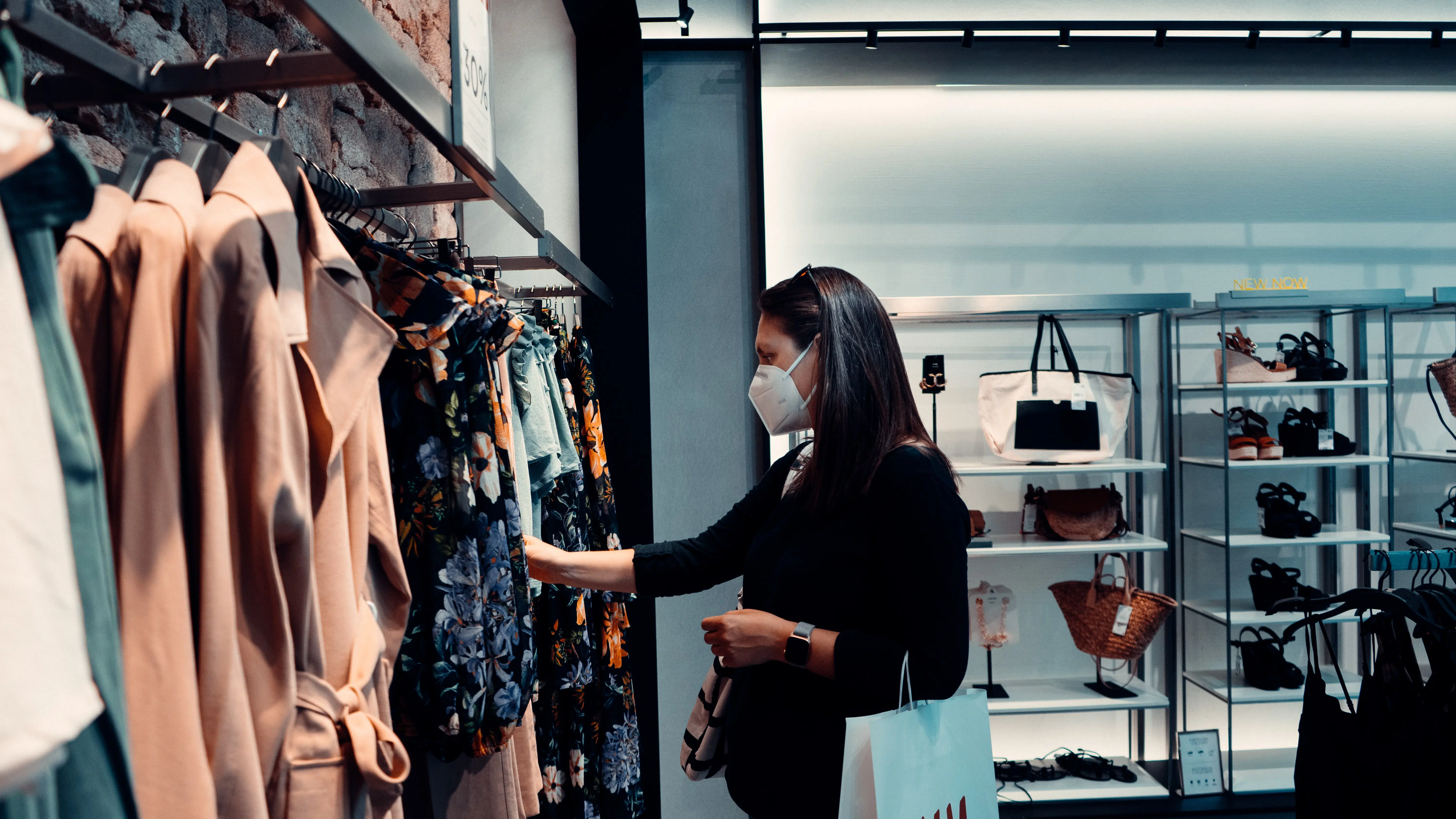 Interior of a store. White woman with black hair and a white face mask looks through a clothing rack.